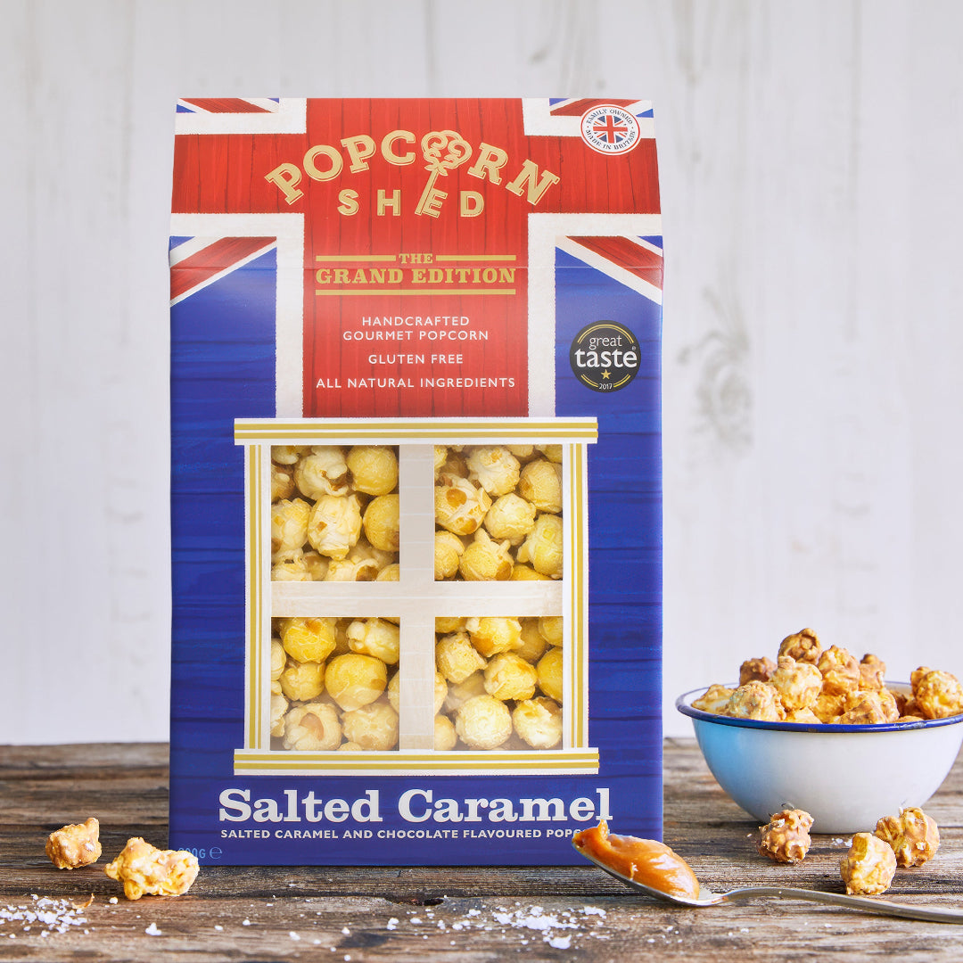 A box of Popcorn Shed Salted Caramel popcorn stands on a rustic wooden surface with scattered caramel popcorn kernels and a small bowl of popcorn to the right. The packaging is blue with red and white Union Jack corner accents and a clear window showing yellow popcorn inside.