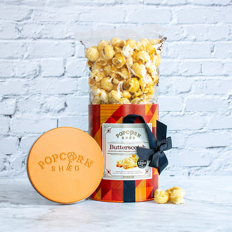 A bag of Popcorn Shred Butterscotch popcorn standing in a clear cylindrical container with a red and orange plaid label, tied with a black ribbon, next to a round peach-colored lid stamped with 'Popcorn Shred' on a white brick background.