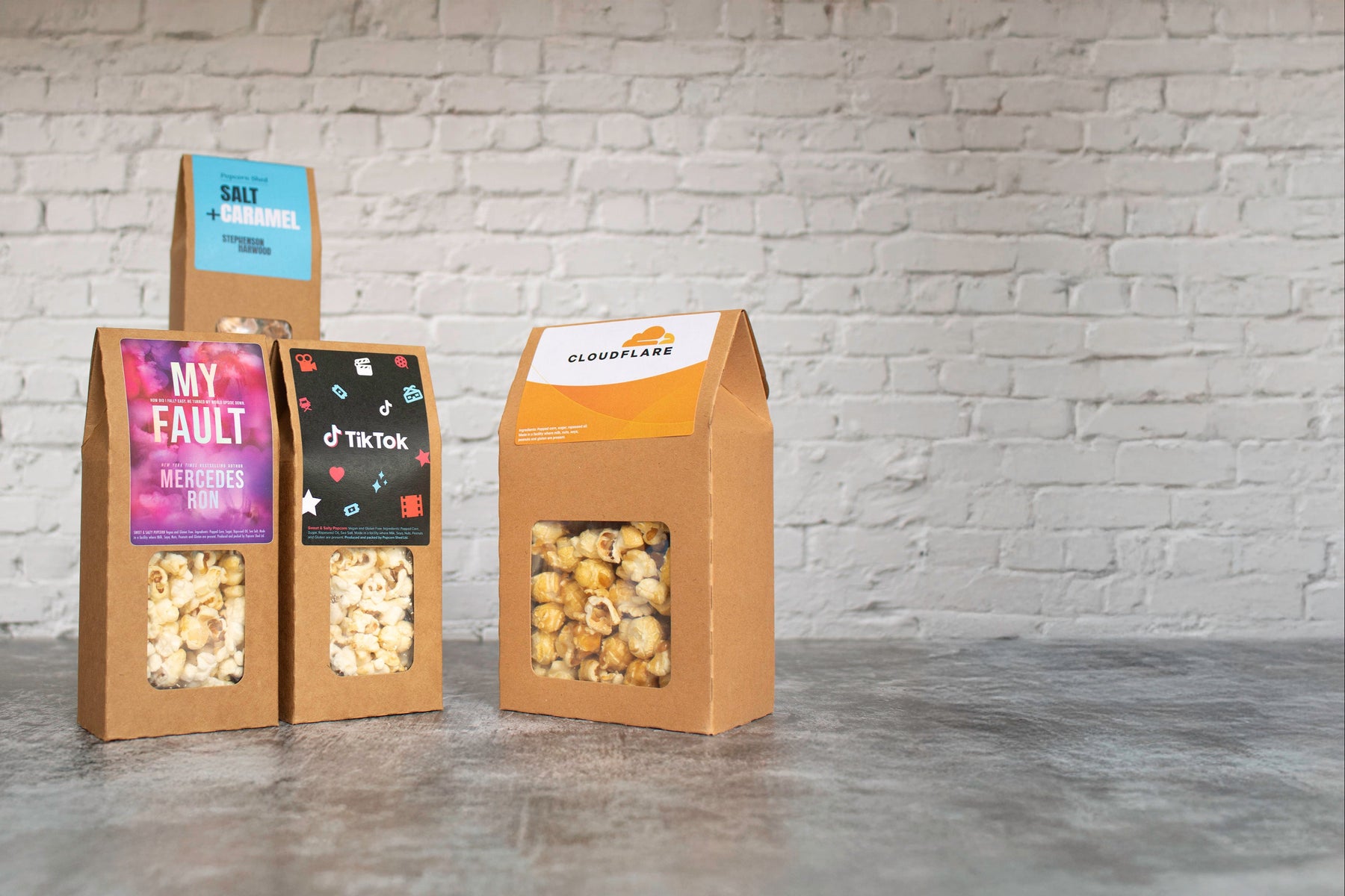 Three cardboard popcorn boxes with different designs and text on a gray surface and brick wall background.